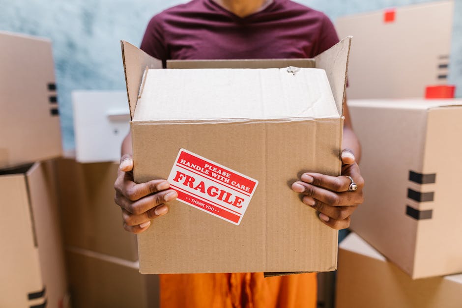 A person holding an open cardboard box with a red and white 'Fragile' handling label, indicating careful packing during a home relocation process. The individual’s hands are visible, showing a wedding ring on the left hand, and they are standing in a room filled with multiple packed moving boxes of various sizes, some with black tape for sealing. The background includes more boxes stacked on a surface, with a doorway and plain wall visible in the setting. The scene is illuminated by natural or soft indoor lighting, emphasizing the packing and moving activity typical of furniture transport and packing services provided by Catford Removals on Brownhill Road, Catford. This visual accurately depicts the careful handling of items during a professional removal, highlighting the importance of proper packing and loading procedures in domestic removals.