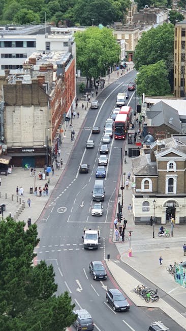 An aerial view of Brownhill Road in Catford showing a busy urban street lined with a mix of residential and commercial buildings, including a red-brick building on the left and a small church-like structure on the right. The street has multiple lanes with cars, vans, and buses moving in both directions, some of which are waiting at a traffic light or junction. Sidewalks are populated with pedestrians, and a few bicycles are parked near the curb. Several mature trees with green foliage line the street, providing shade and greenery to the scene. The environment appears well-lit with natural daylight, capturing a typical city traffic and pedestrian activity, relevant to the home relocation and furniture transport context for Catford Removals, with trucks and moving equipment possibly involved in the background.