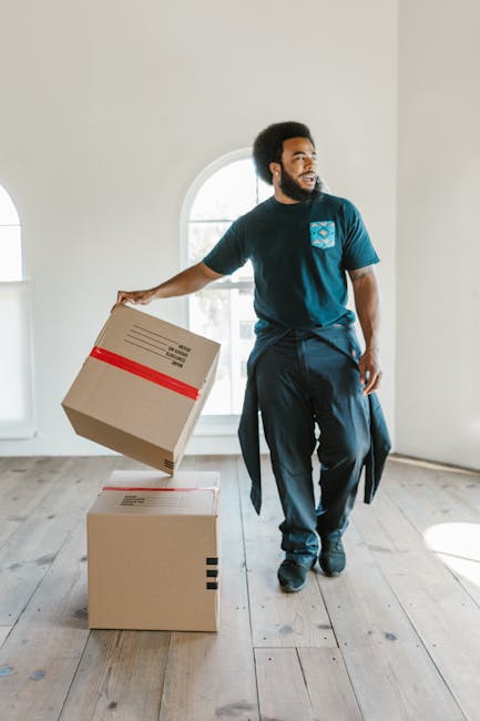 A professional mover from Catford Removals, wearing a dark blue T-shirt with a company logo and dark trousers, is inside a bright, spacious room with white walls and large arched windows. He is lifting a cardboard box wrapped with red packing tape, preparing for a home relocation or furniture transport. On the wooden floor, there are two similar cardboard boxes, one stacked on top of the other, indicating an organized packing process. The room is well-lit with natural light coming through the windows, and the mover is engaged in a lifting and carrying action typical of packing and moving activities. This scene illustrates the careful handling of boxes and furniture during a professional removals service, emphasizing the logistics involved in home relocation, with the environment showing a clean and empty interior ready for furniture loading or unloading.