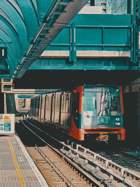 A modern underground train with a white front and blue and orange accents is positioned on railway tracks beneath a green metal bridge or structure. The train's windows are tinted, and its door is closed. To the left, part of a train platform is visible, featuring tactile paving strips and safety signage. The scene is illuminated by natural daylight, highlighting the urban environment typical of a city train station. This image captures the process of moving or transportation within public transport infrastructure, relevant to home relocation or furniture transport services offered by Catford Removals, especially around the area near Catford Bridge station.