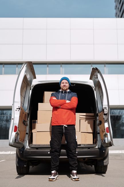 A man standing with arms crossed in front of an open cargo van filled with neatly stacked cardboard boxes, ready for home relocation or furniture transport. The van, parked outdoors outside a modern building with a white facade and large windows, is part of a scheduled move handled by Catford Removals. The man is dressed casually in a blue beanie, red and navy jacket, dark trousers, and sneakers, indicating a professional loading process. Inside the van, the boxes are arranged on a loading area with visible details of packaging materials, and the vehicle appears prepared for transport. The scene captures a moment of packing and moving logistics, highlighting the practical aspect of house removals or furniture transport services, supported by equipment like the van and the organized packing materials.