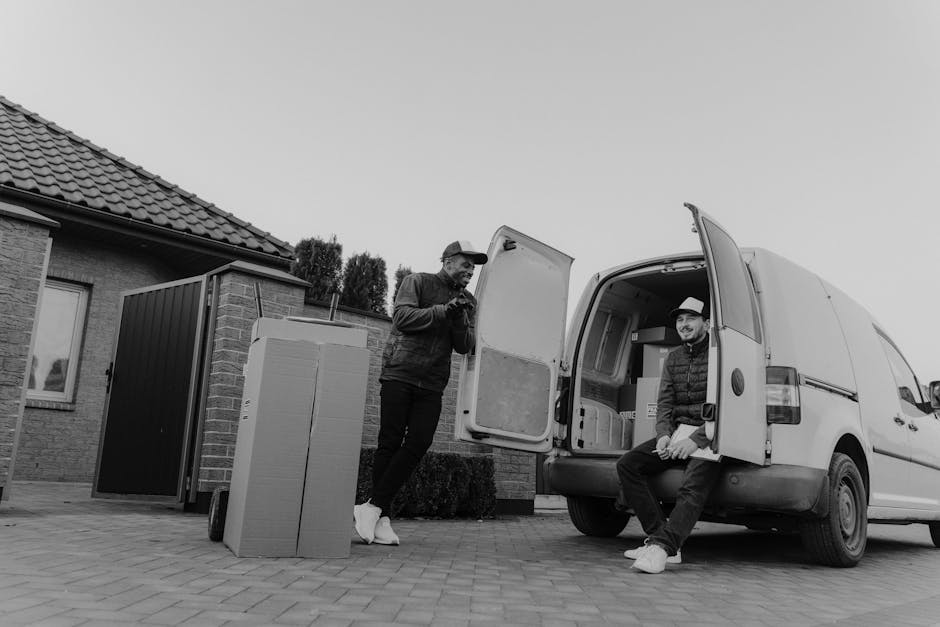 Two men involved in a home relocation process are positioned near a white moving van parked outdoors on a paved driveway in front of a residential building with a tiled roof. One man, standing outside the van, is dressed in dark clothing and wearing a cap, looking at his phone or a small device. The second man is seated on the edge of the van’s open rear door, wearing a light-colored hat, dark jacket, and light trousers, smiling and facing the camera. In front of the van, there are several large cardboard boxes and a flat-panel item, possibly a piece of furniture or appliance packaging, resting on a trolley with wheels. The scene occurs in natural daylight, with the interior of the van visible showing empty space prepared for loading items for a furniture transport or moving service. The setup is typical of a professional removals operation by Catford Removals, illustrating the packing and loading process involved in house removals or house clearance, with visible focus on the vehicle, packing materials, and the teamwork involved in furniture transport and logistics.