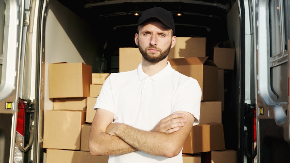 A man standing with arms crossed in front of an open cargo van filled with neatly stacked cardboard boxes, ready for home relocation or furniture transport. The van, parked outdoors outside a modern building with a white facade and large windows, is part of a scheduled move handled by Catford Removals. The man is dressed casually in a blue beanie, red and navy jacket, dark trousers, and sneakers, indicating a professional loading process. Inside the van, the boxes are arranged on a loading area with visible details of packaging materials, and the vehicle appears prepared for transport. The scene captures a moment of packing and moving logistics, highlighting the practical aspect of house removals or furniture transport services, supported by equipment like the van and the organized packing materials.