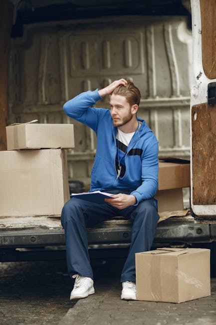 A young man with light brown hair, dressed in a blue hoodie, white T-shirt, and dark blue trousers, is sitting on the edge of a moving van’s loading area. He is holding a clipboard and appears to be